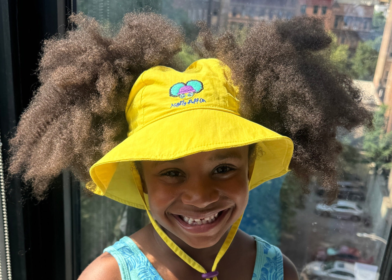 A smiling kid wearing the yellow Mighty Puff Hat, afro puffs flowing through the back opening