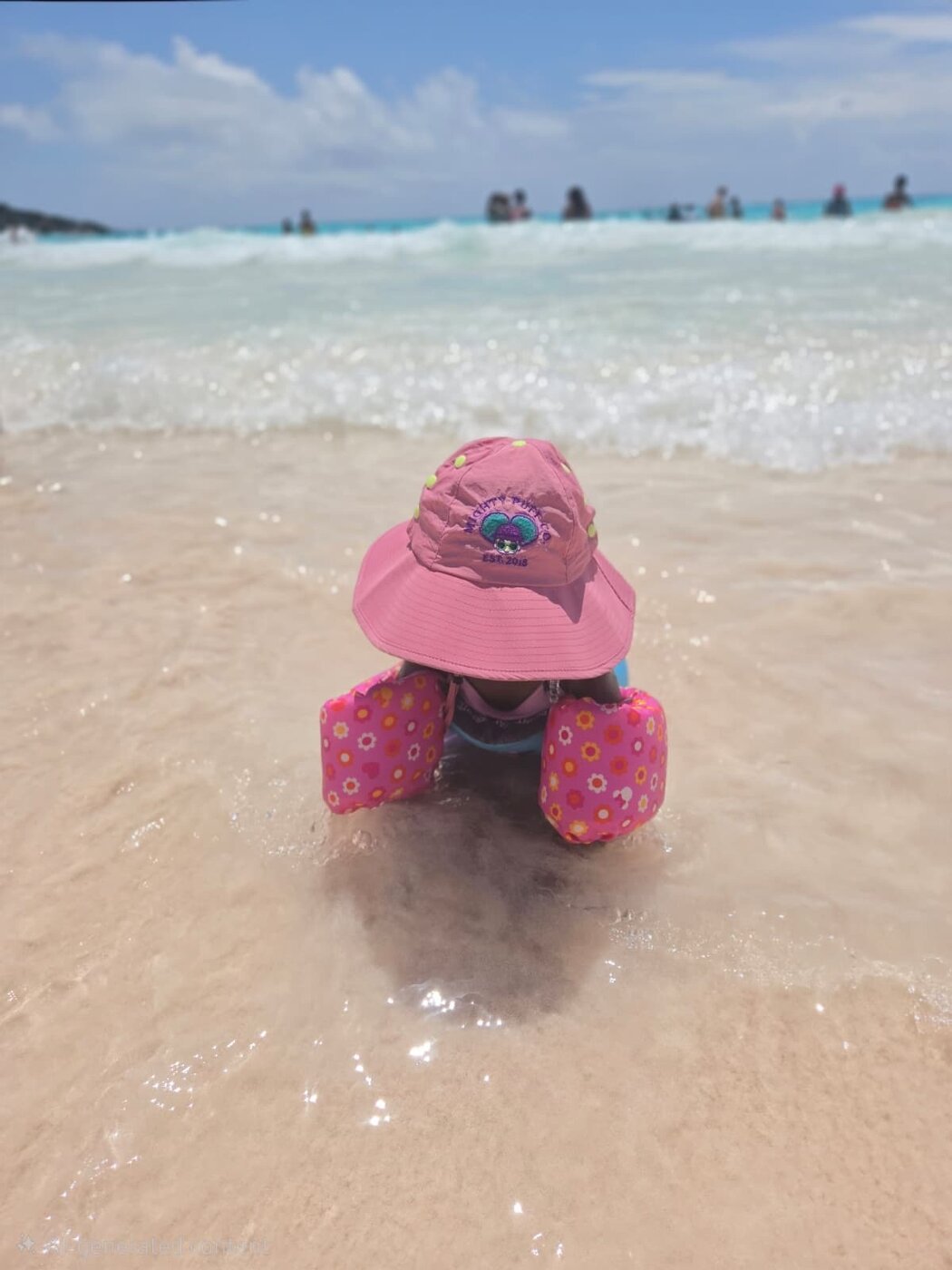A little one in the pink hat sitting in the ocean surf with floaties