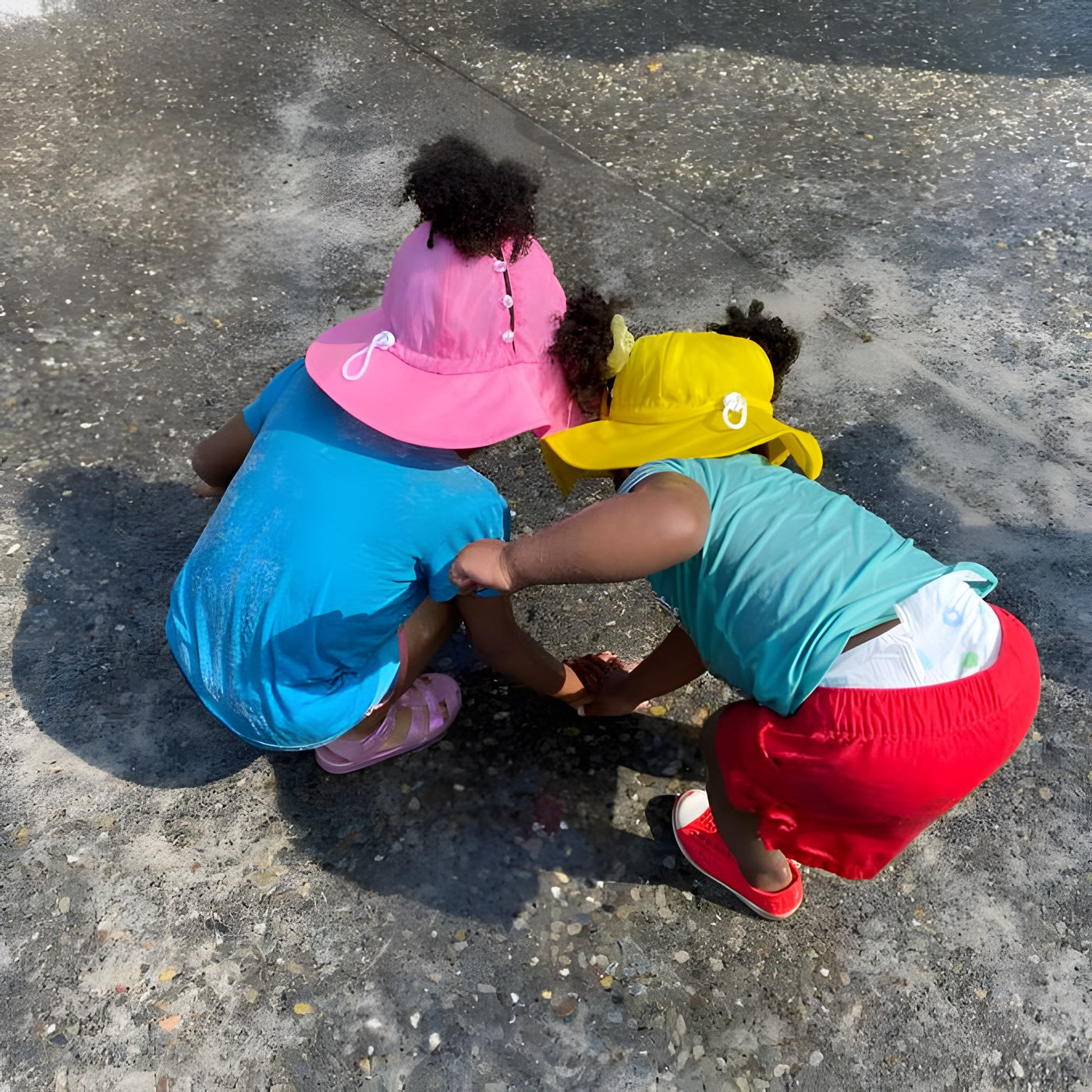 Two toddlers in pink and yellow Mighty Puff hats crouched over a puddle